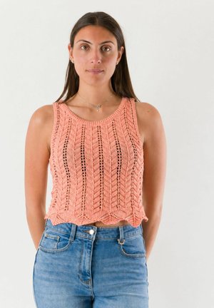 Donna con lunghi capelli scuri che indossa un top rosa lavorato a maglia senza maniche e jeans blu, in piedi davanti a uno sfondo chiaro e semplice.