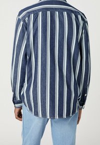 Man i en marinblå och vit vertikalt randig långärmad skjorta och ljusblå jeans, sedd bakifrån mot en neutral bakgrund.