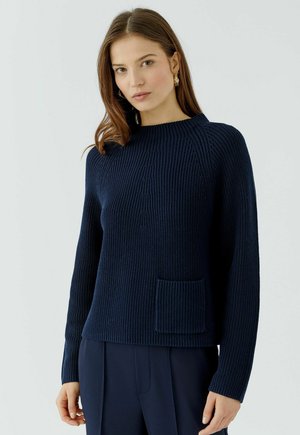 Vrouw met recht bruin haar, gouden oorbellen, een donkerblauwe geribde trui met voorzak en donkerblauwe broek tegen een effen achtergrond.