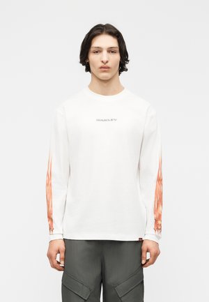 Νεαρός με μεσαίου μήκους σκούρα μαλλιά φοράει λευκό πουκάμισο με μακριά μανίκια και πορτοκαλί φλόγες στα μανίκια και γκρι παντελόνι cargo, κοιτάζοντας μπροστά.