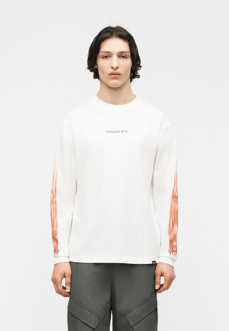 Νεαρός με μεσαίου μήκους σκούρα μαλλιά φοράει λευκό πουκάμισο με μακριά μανίκια και πορτοκαλί φλόγες στα μανίκια και γκρι παντελόνι cargo, κοιτάζοντας μπροστά.