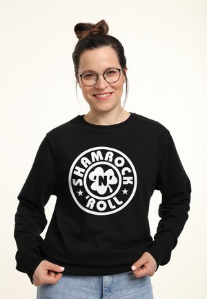 Smilende kvinde med briller og hår sat op i en knold iført en sort sweatshirt med teksten "Shamrock n Roll" og et kløvergrafik, lys baggrund.
