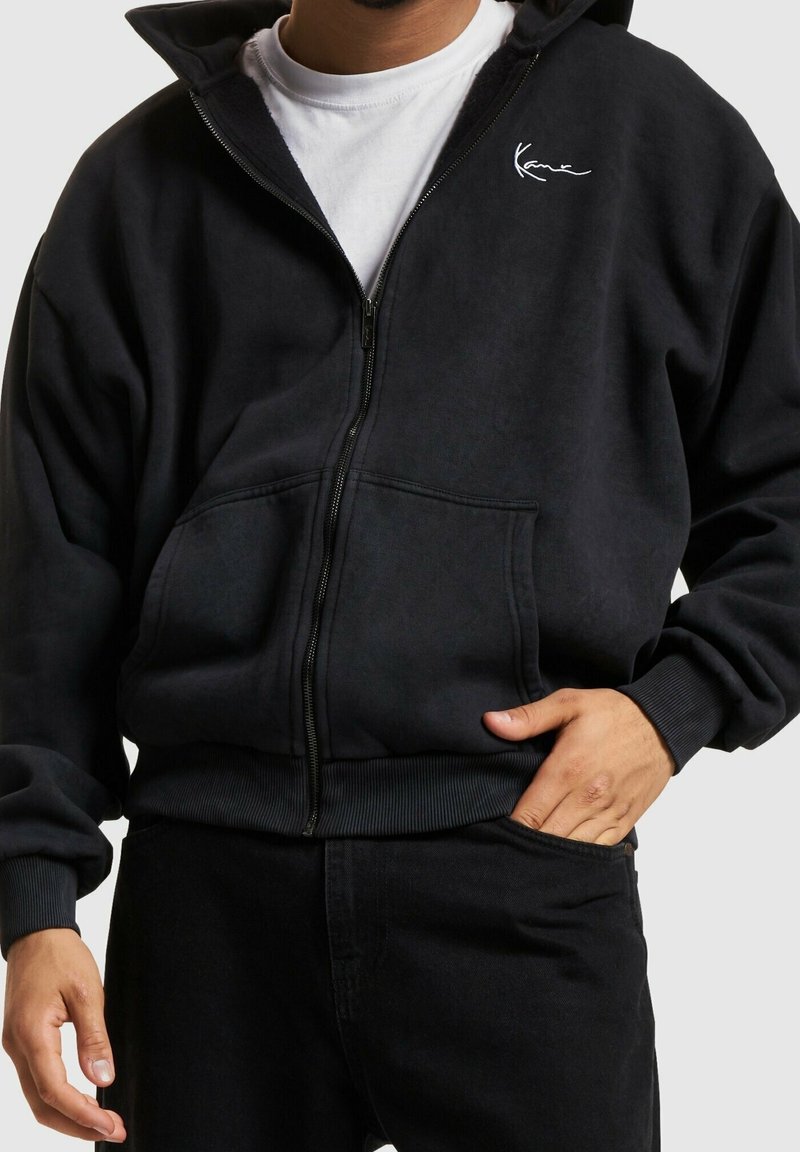 Homme portant un sweat à capuche noir zippé avec un petit logo blanc sur la poitrine et un pantalon noir, la main dans la poche.