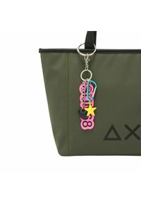 Zielona torba na ramię z czarnymi akcentami, ozdobiona różnorodnymi kolorowymi breloczkami w zabawnych kształtach, w tym różowymi, żółtymi i czarnymi.