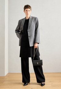 Blazer de lana gris con dos botones, combinado con un vestido negro y pantalones negros de pierna ancha. Sosteniendo un pequeño bolso negro con cadena.
