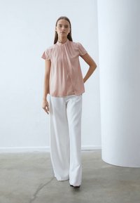 Kvinne i kortermet rosa bluse og vide hvite bukser, stående med den ene hånden på hoften mot en minimalistisk hvit bakgrunn.