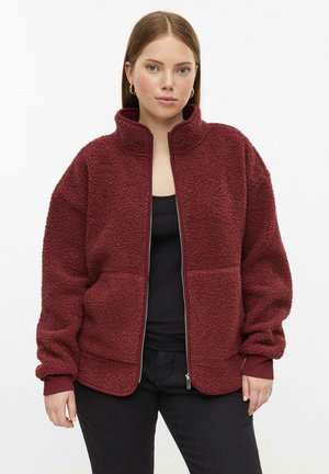 Frau trägt eine burgunderrote Fleecejacke mit Reißverschluss über einem schwarzen Top und schwarzen Hosen, steht vor einem einfachen weißen Hintergrund.