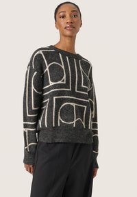 Stickad tröja med svart bas och kontrasterande vita geometriska mönster. Har en avslappnad passform och ribbat fåll och ärmar.
