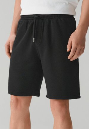 Férfi fekete, combközépig érő zsinóros rövidnadrágban és fehér ingben, kezeit lazán oldalt tartva, sima háttér előtt áll.