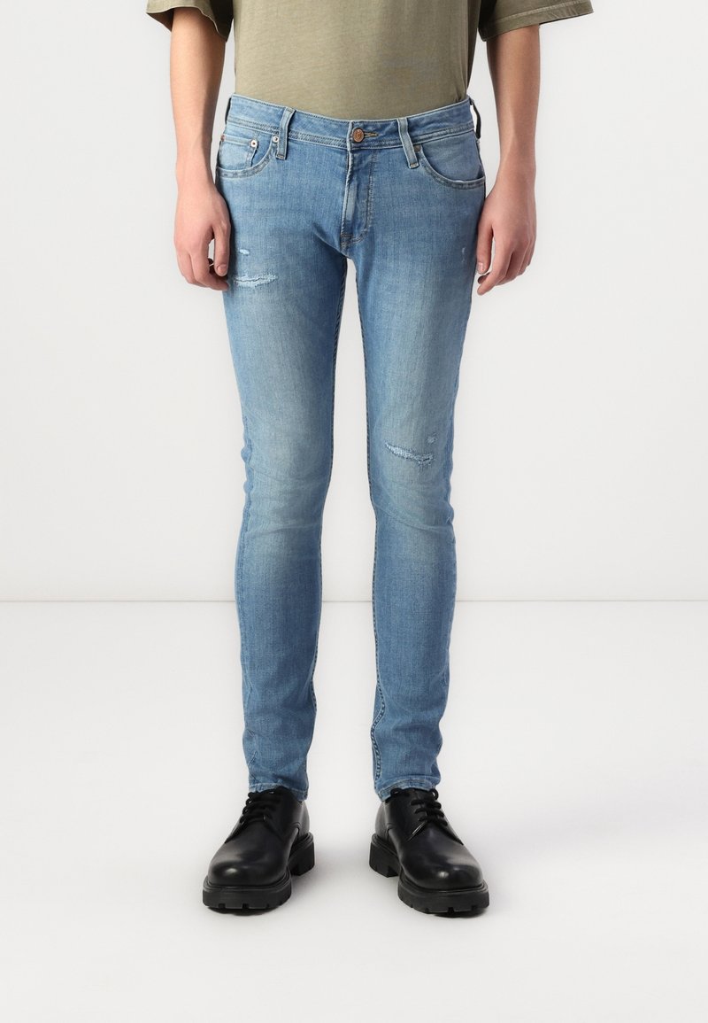 Mann trägt hellblaue Slim-Fit-Jeans mit leichten Gebrauchsspuren und schwarze Schnürschuhe, steht vor einem schlichten weißen Hintergrund.