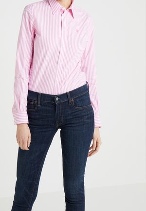 Vrouw met een roze-wit gestreept overhemd met knopen, ingestopt in donkerblauwe slim-fit jeans, op een witte achtergrond.