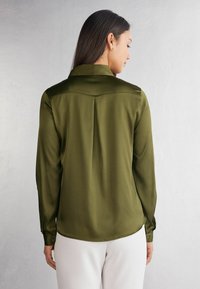 Blusa verde oliva a maniche lunghe con colletto, dettaglio con piega sul retro, texture liscia e polsini con bottoni, abbinata a pantaloni di colore chiaro.