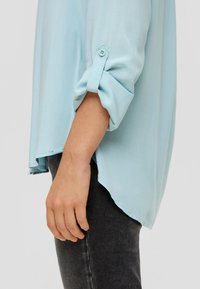 Helles blaues Button-Down-Hemd mit hochgekrempelten Ärmeln, aus weichem Stoff. Verfügt über einen abgerundeten Saum und einen dezenten Glanz, kombiniert mit dunklen Jeans.