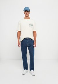 Mann står med fronten vendt mot oss, iført en lys beige "Birding Club" T-skjorte, blå jeans, hvite sneakers og en lys blå denim caps mot en enkel hvit vegg.