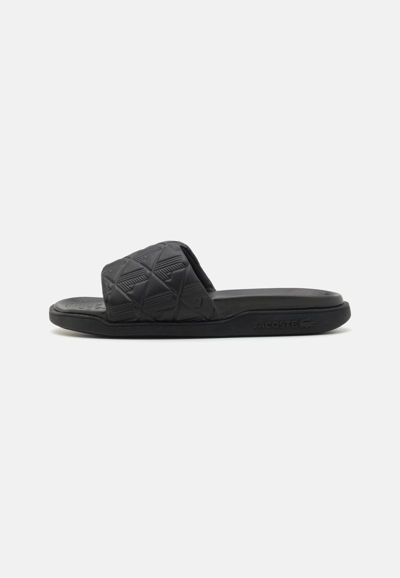 Sandale noire à enfiler avec une large bride texturée et une semelle plate, présentant un motif en relief subtil et le logo Lacoste sur le côté.