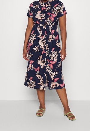 Vrouw draagt een midi-jurk in marineblauw met roze bloemen- en vlinderdetails, groene sandalen met open teen, staand tegen een effen witte achtergrond.