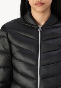 Veste rembourrée noire avec un matelassage en chevrons, finition brillante, col haut et fermeture éclair argentée. Texture lisse, sans logos ni motifs visibles.