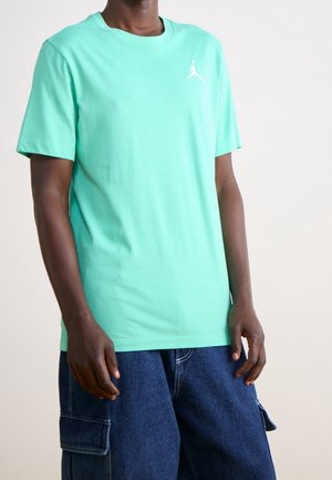 Osoba nosząca jasnoturkusowy t-shirt z krótkim rękawem z małym białym logo oraz ciemnoniebieskie szorty cargo z denimu.