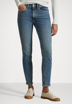 Magas derekú, slim-fit farmernadrág világoskék denim anyagból, öt zsebbel, gombos záródással és minimális kopással. Bézs vászoncipővel párosítva.