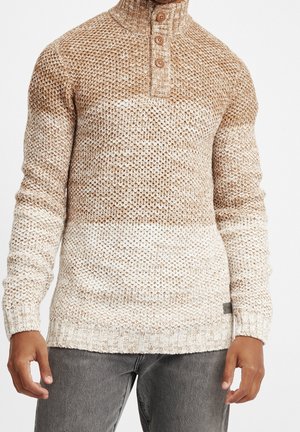 Homme portant un pull en maille beige et blanc avec trois boutons au col montant, associé à un jean gris foncé.