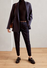 Navy blazer and fitted black turtleneck paired with tailored navy trousers. Accessories include a brown belt and leather messenger bag. Black loafers.