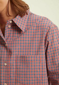Chemise à carreaux rouges et bleus en tissu de coton tissé, avec un col boutonné et une poche poitrine avec des détails de couture.