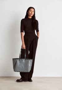 Femme portant un haut noir et un pantalon large tenant un grand sac fourre-tout à motifs, debout contre un mur blanc uni.