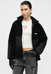 Μαύρο μπουφάν από φλις και νάιλον με φερμουάρ και ψηλό γιακά, με λευκό λογότυπο. Φορεμένο πάνω από ένα λευκό πουκάμισο με ροζ καρδιά. Μαύρα γάντια.