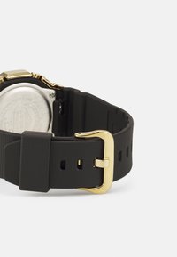 Bracelet de montre en caoutchouc noir avec une boucle en métal doré, présentant une surface texturée et plusieurs trous de réglage, fixé à un dos de montre en argent.