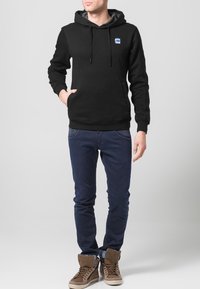 Felpa nera con tasca frontale e coulisse regolabili, abbinata a jeans blu scuro. Presenta un piccolo logo blu sul petto.