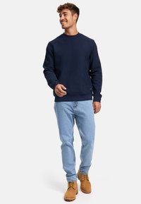 Sudadera texturizada azul marino con cuello redondo, combinada con jeans azul claro y botas de trabajo color marrón claro. El modelo adopta una pose casual.