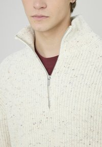 Pull beige clair en tricot avec col zippé et variations de texture. Présente des éclats multicolores dans tout le tissu.