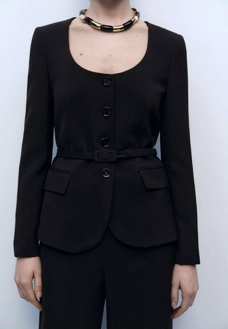 Femme portant un blazer noir cintré à quatre boutons, taille ceinturée, poches à rabat, et un collier épais noir et doré.