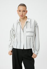 Cropped button-up shirt in white fabric with black vertical stripes, featuring a chest pocket and loose-fitting long sleeves.