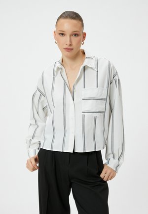 Cropped button-up shirt in white fabric with black vertical stripes, featuring a chest pocket and loose-fitting long sleeves.