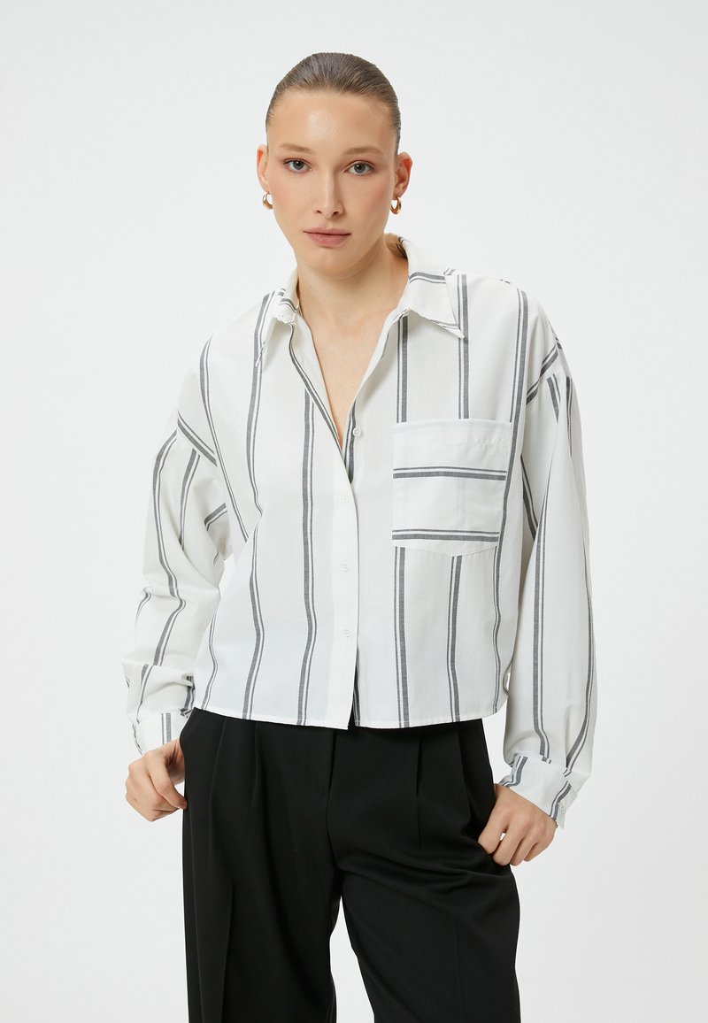 Cropped button-up shirt in white fabric with black vertical stripes, featuring a chest pocket and loose-fitting long sleeves.