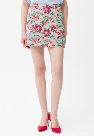 Kvinde iført en beige mininederdel med røde og grønne blomster, kombineret med røde højhælede sko og en lyseblå bluse.