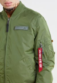 Olīvu zaļas krāsas bombers jaciņa ar rāvējslēdzēju, ribota apkakle un vairākām kabatām. Uz tās ir piestiprināta sarkana etiķete ar uzrakstu "REMOVE BEFORE FLIGHT".