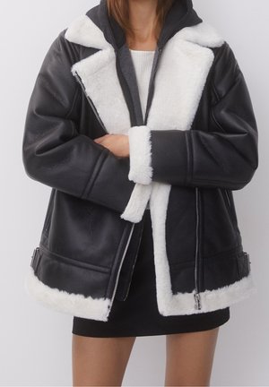 Personne portant une veste en cuir noire avec une doublure et des poignets en shearling blanc, debout, les bras croisés sur un haut blanc et une jupe noire.