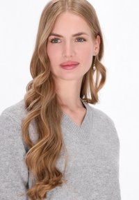 Szürke, V nyakú pulóver puha kötött anyagból. A modellen hosszú, hullámos szőke haj van, és gyöngy fülbevalót visel. Egyszerű fehér háttér.