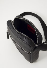 Borsa a tracolla in pelle nera con finitura testurizzata, dotata di chiusura con zip argentata, logo impresso e tracolla regolabile. L'interno mostra un'etichetta rossa.