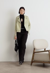 Jachetă de biker din piele verde deschis cu accesorii argintii, pulover negru cu gât și pantaloni. Accesorizată cu o geantă mică neagră și cizme cu toc.