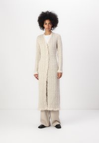 Gros cardigan beige en tissu texturé, avec fermeture à bouton et bords effilochés. Porté sur une chemise blanche ; le modèle pose sur fond uni.