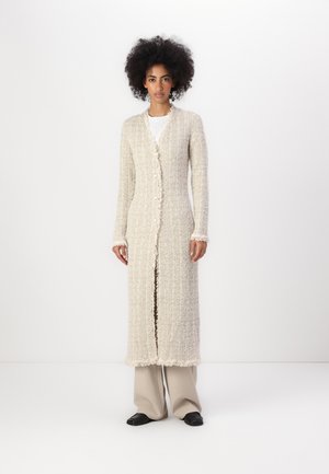 Cardigan lungo beige con tessuto texturizzato, chiusura con bottoni e bordi sfrangiati. Indossato sopra una camicia bianca; il modello posa su uno sfondo semplice.