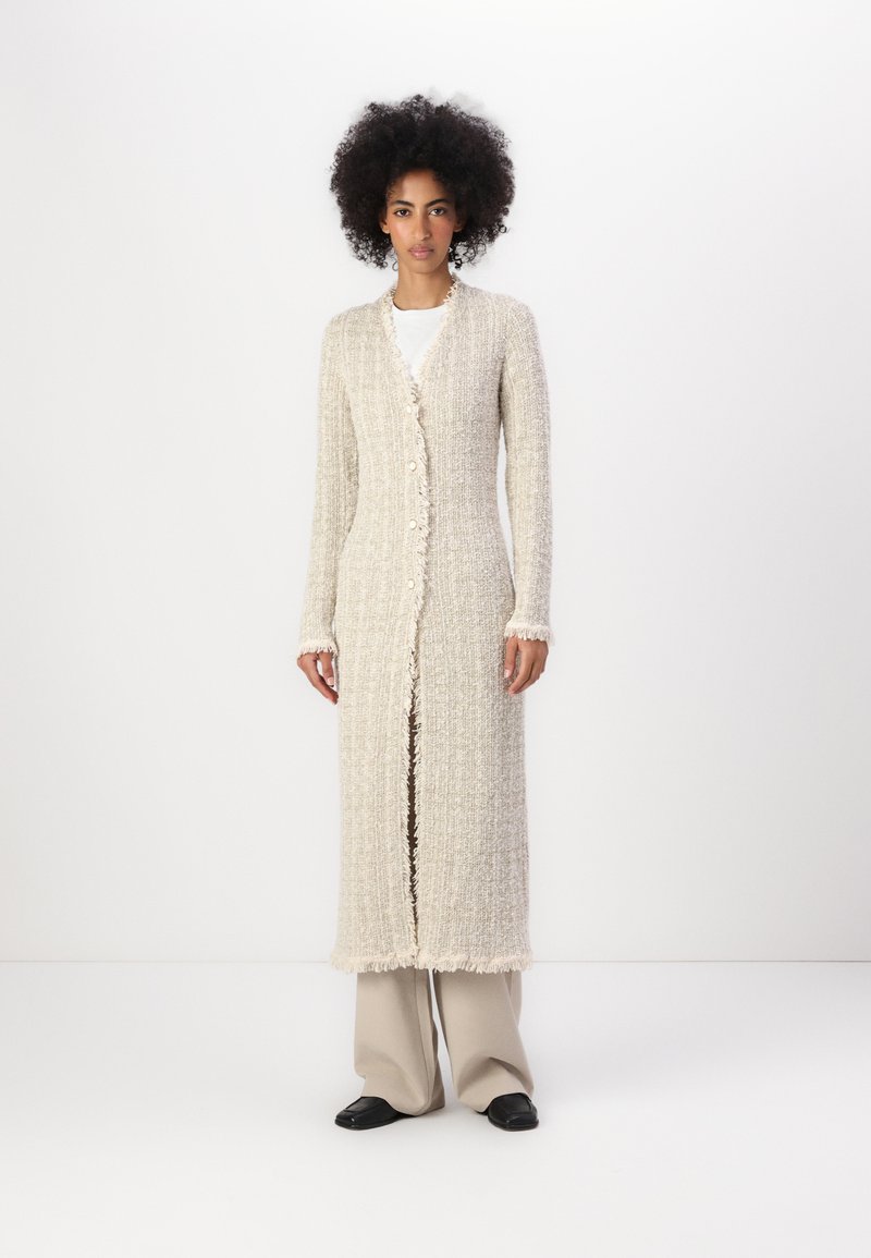 Gros cardigan beige en tissu texturé, avec fermeture à bouton et bords effilochés. Porté sur une chemise blanche ; le modèle pose sur fond uni.