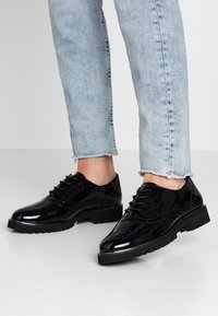 Scarpe brogue nere lucide con lacci indossate con jeans azzurri con orlo sfrangiato, in piedi su un pavimento bianco liscio.