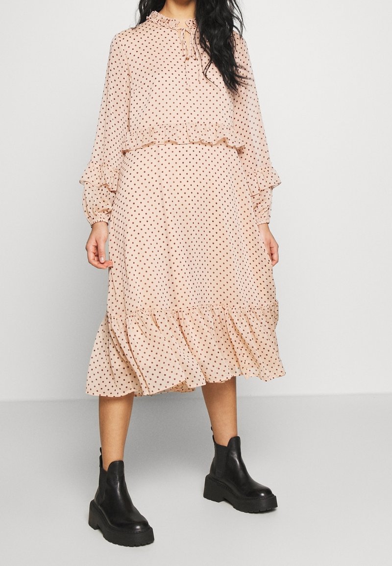 Femme portant une robe midi rose clair à pois avec des volants et des manches longues, associée à des bottines noires épaisses.