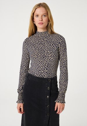 Vrouw met lang blond haar draagt een marineblauw langemouwen top met witte stippen en een hoge hals, gecombineerd met een zwarte rok met knopen.