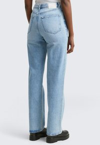 Person som bär ljusblå högmidjade raka jeans och svarta kraftiga stövlar, visad bakifrån mot en enkel bakgrund.