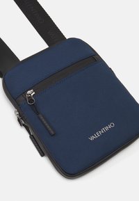 Plavi najlon torbica preko tijela s crnim zatvaračem, glatkim detaljima i srebrnim "Valentino" logotipom sprijeda. Kompaktni pravokutni oblik.